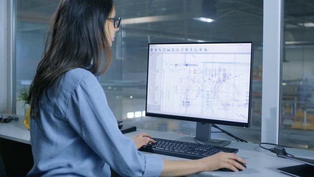 Female Industrial Engineer Working With Technical Blueprints on Her Personal Computer. Inside of the Factory is Seen From Her Office Window. Shot on RED EPIC-W 8K Helium Cinema Camera.