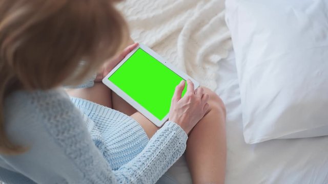 Young Woman in blue sweater sitting on the bed uses Tablet PC with pre-keyed green screen. Few types of gestures - scrolling up and down, tapping, zoom in and out. Perfect for screen compositing