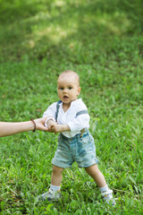 A little boy with dark eyes in a white shirt and denim shorts, trying to make the first steps on the green grass