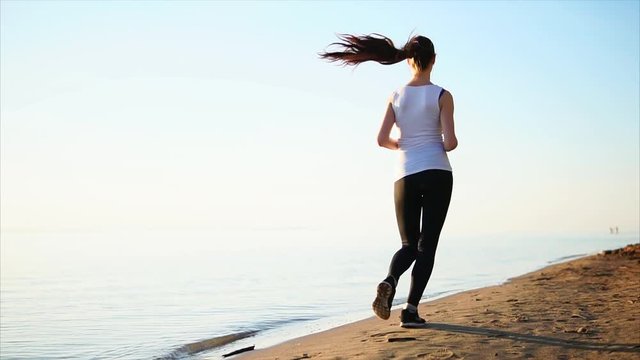Young woman running on the sand beach near the sea. Female jogging view from the back.