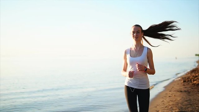 Happy smiling sport woman jogging on the sand beach near the sea. Female listen to the music and wearing a smart watches while she running.