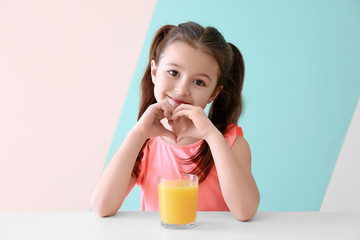 Cute little girl with glass of juice sitting at table, on color background