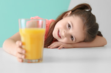 Cute little girl with glass of juice sitting at table, on color background