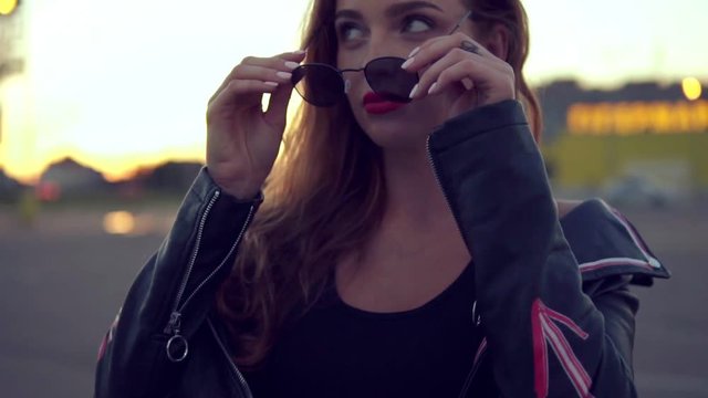 Young charming woman with red lips and long hair in leather jacket wears sunglasses