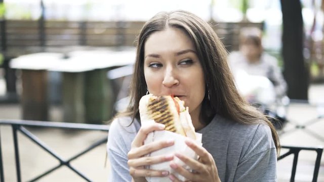 Beautiful Woman Wearing A Gray T Shirt Is Sitting In A Cafe Outside And Enjoying A Large And Tasty Sandwich. Handheld Real Time Close Up Shot