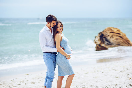 Courageous and handsome man in a white shirt and blue trousers  hugs her beautiful pregnant wife in a blue dress against the background of the rocks and the sea.