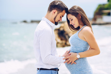 Loving couple on the beach touching pregnant woman's belly with love and care on the background of rocks and sea in sunny day.