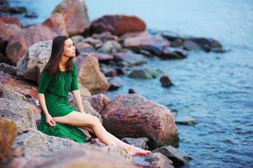 Cute girl in green dress sitting barefoot on a large rock on the seashore and looks into the distance.