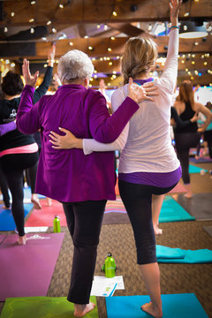 Vertical photo of grandmother and granddaughter in group yoga practice with colorful mats and festive lighting. Soft focus background with room for text/copy. 