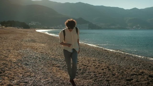 Young man dances on beach in summer day. He rhythmically and expressively moves with light music on picturesque coast of Montenegro in sunny weather. Curly-haired brunette in sunglasses dressed in