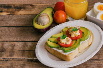 Open sandwich style for breakfast or lunch. Sandwich spread with cream cheese, avocado, tomato and boiled eggs on white plate. Avocado open sandwich style serve with orange juice on rustic wood table.