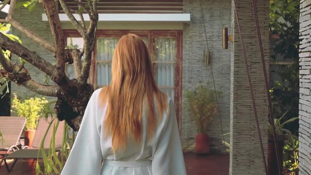 Young woman walking in bathrobe towards door of villa, view from her back, tracking shot