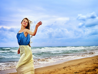 Woman on beach in sunset on sand. Teenager girl on sea shore. Summer style outfit sitting near ocean. Girl on uninhabited island. Girl wearing fashionable dress waving goodbye hand.