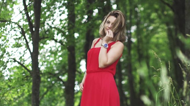 Portrait of a young woman in a red summer dress against a background of green trees. slow motion
