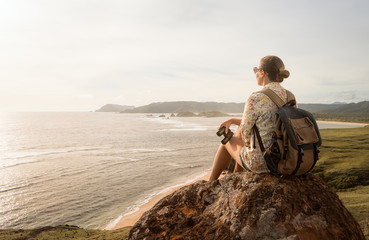 Woman traveller with backpack and binoculars in hands enjoying view coast on sunset