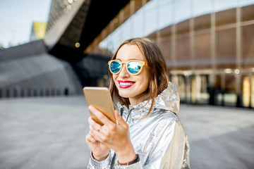 Lifestyle portrait of a stylish woman in silver jacket talking with phone outdoors on the modern architectural background