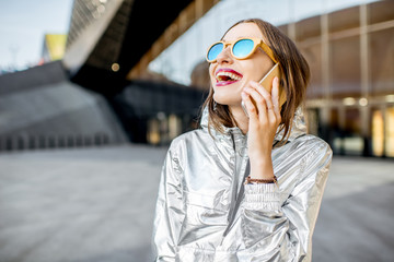 Lifestyle portrait of a stylish woman in silver jacket talking with phone outdoors on the modern...