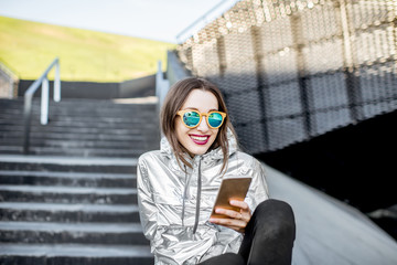 Lifestyle portrait of a stylish woman in silver jacket sitting with phone outdoors in the modern architectural environment