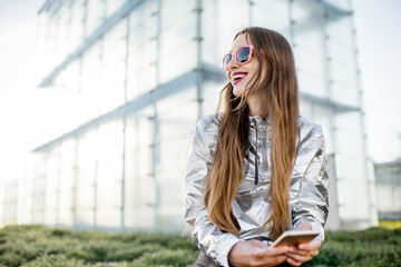 Lifestyle portrait of a stylish woman in silver jacket sitting outdoors at the modern architectural environment