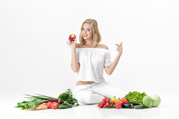 Beautiful blond woman in white clothes and lots of fresh vegetables on white background. girl is eating nectarine