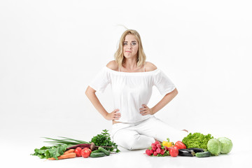 Beautiful upset blond woman in white clothes and lots of fresh vegetables on white background. diet