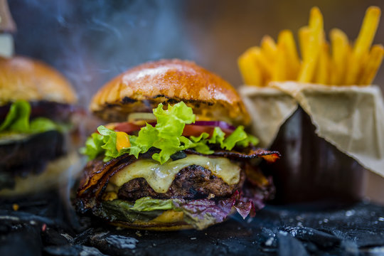 Tasty smoked grilled and glazed beef burger with lettuce, cheese and bacon served with french fries on wooden table with copyspace, smoke mesquite timber wood in background.