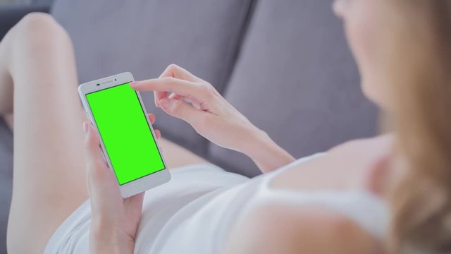 Young Woman in white top laying on couch uses SmartPhone with pre-keyed green screen. Few types of gestures - scrolling up and down, tapping, zoom in and out. Perfect for screen compositing