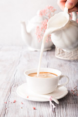 White cup of coffee with pink flowers on old white wooden background with copy space. Milk is poured into a cup.