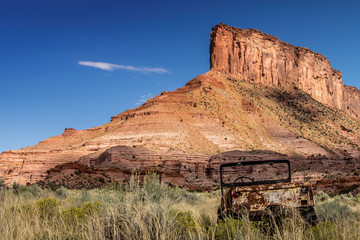 abandoned rusty jeep in a field at the base of a red rock mesa with a bright blue sky