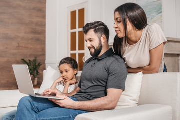 Fototapeta premium multiethnic family sitting on sofa and using laptop together