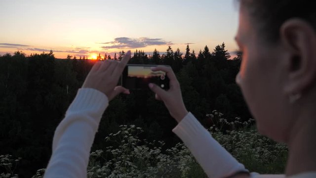 The girl shoots and takes pictures of the sunset using a smartphone.