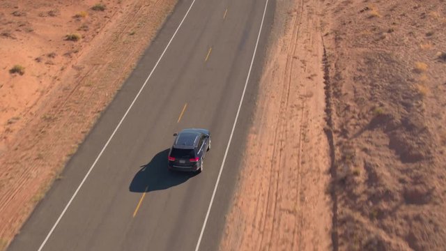 AERIAL CLOSE UP: Flying above the black SUV car driving along an empty road trough sandstone desert in Utah flatlands, USA. People on road trip traveling on road trough desert landscape on sunny day.