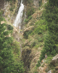 A man sits on a rock near a waterfall and looks up