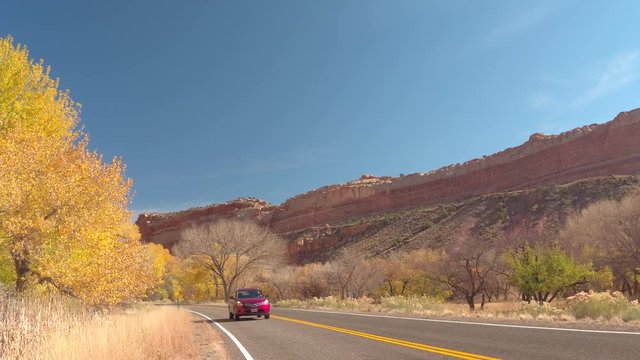 CLOSE UP: Red Car Driving Along An Empty Road Through Red Rock Canyon Past Stunning Bright Yellow Trees With Turning Leaves On Sunny Autumn Day. Road Trip Trough Capitol Reef Park In Lush Fall