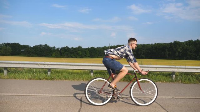Young handsome man riding at vintage bicycle in the country road. Sporty guy cycling at the track. Male cyclist riding fixed gear bike at highway. Healthy active lifestyle Slow motion