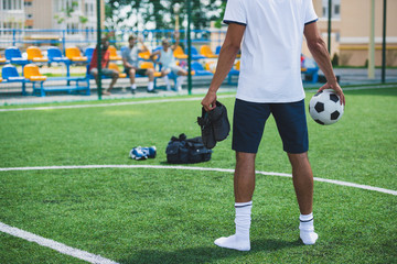 Obraz premium back view of soccer player with ball and football boots in hands standing on pitch