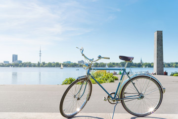 vintage blue bicycle parked at lakeshore of Alster Lake in Hamburg, Germany under beautiful clear blue summer sky with cityscape in blurred background - Powered by Adobe