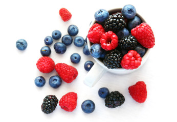 Ripe blueberries, raspberries and blackberries in a white cup and next to it.