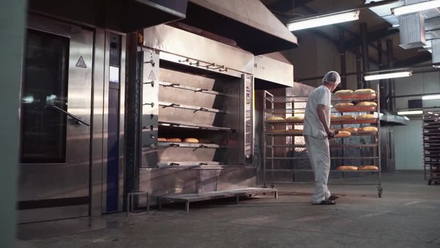 A male baker in a white uniform takes out of the oven just baked bread, places it on the tray. Close up view of ready-made loaves of bred on the cooling rack. Manufacturing process, bakery equipment.