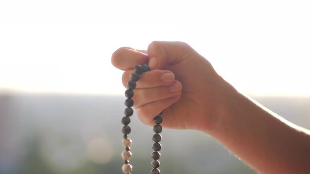 Female lit hand close up on sunset or sunrise, counts Malas strands of gemstones beads used for keeping count during mantra meditations. Woman sits on top above city. Slow motion.