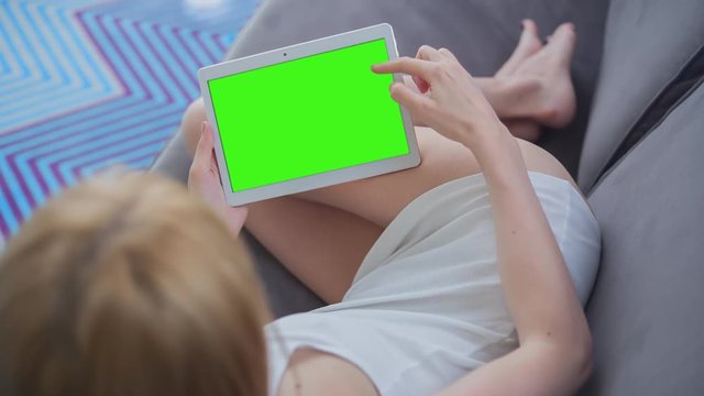Young Woman in white top laying on couch uses Tablet PC with pre-keyed green screen. Few types of gestures - scrolling up and down, tapping, zoom in and out. Perfect for screen compositing