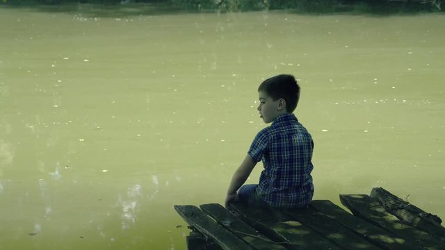 Dreamy boy sitting on wooden pier at pond. Little kid thinks and looks at pond. Dream positive atmosphere, sunny day.