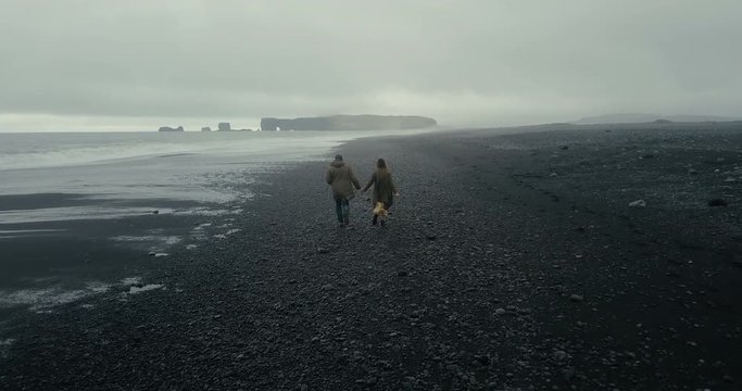 Back aerial view of young hipster couple walking on the black volcanic beach in Iceland. Man and woman enjoy the sea.