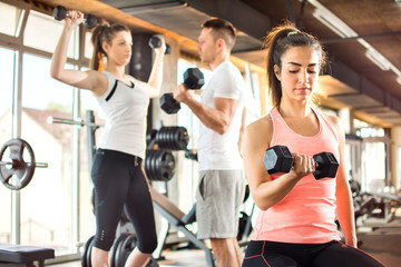 Young woman lifting dumbbells at the gym with friend in the background.