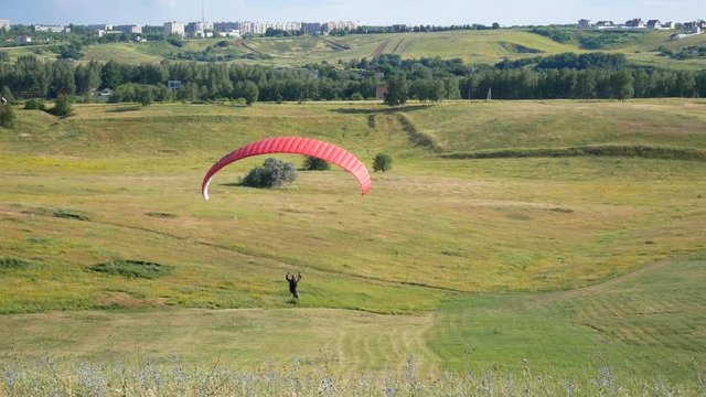 Person Fly On Paraglider On Background Of Green Grass On Sunny Day. Man Paragliding. Glider Moves Smoothly Through The Air. Paragliders Flying An Extreme Sports Event On A Paraglider
