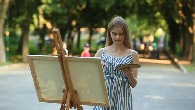 Beautiful girl draws a picture in the park using a palette with paints and a spatula. Easel and canvas with a picture. Summer is a sunny day, sunset.
