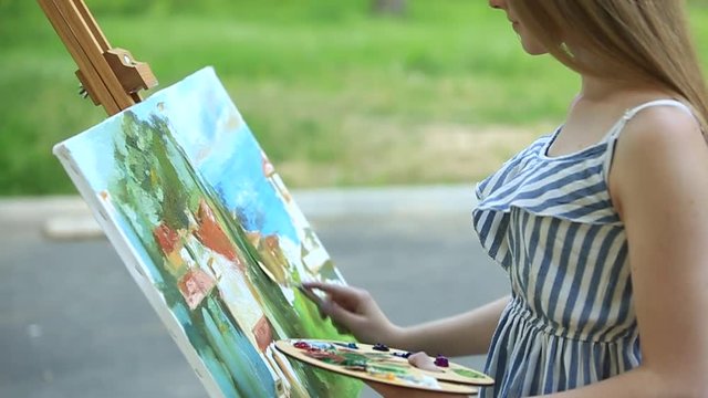 Beautiful girl draws a picture in the park using a palette with paints and a spatula. Easel and canvas with a picture. Summer is a sunny day, sunset.