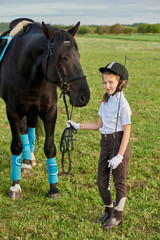 Pretty little girl jockey communicating with her black horse in professional outfit