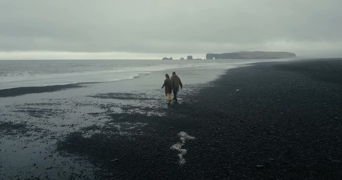Aerial view of young stylish couple walking on the black volcanic beach in Iceland. Man and woman running from the wave.