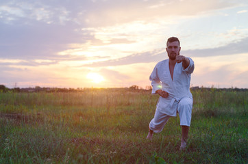 Naklejka na ściany i meble A man in a kimono trains karate at sunset, a copy of space. Beautiful landscape: field and sky with sunset.
